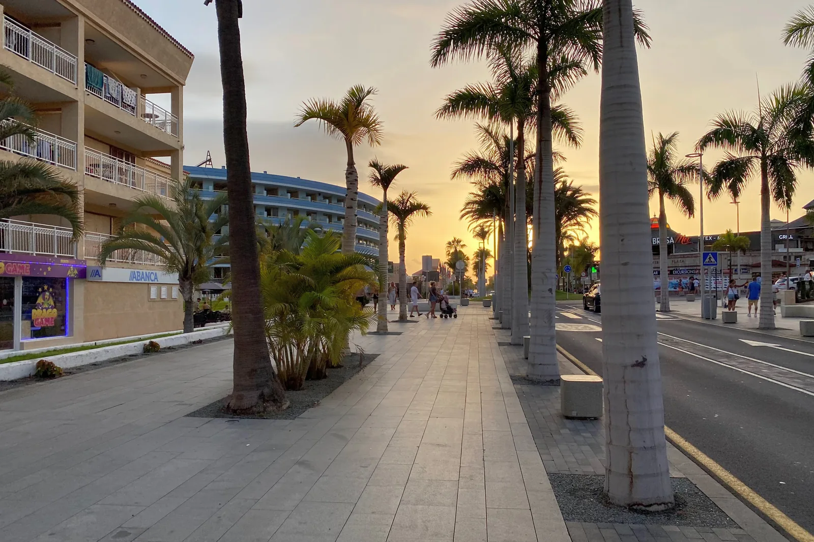 A pedestrian avenue lined with palms in Playa de las Américas at sunset, with apartment buildings on the left, a road on the right and warm light between the trunks