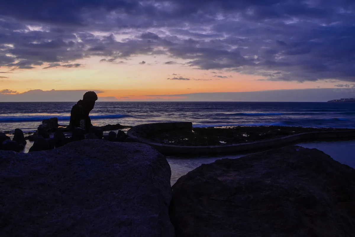 The sun setting over the Atlantic at Playa de las Américas, with an orange and purple sky, a line of surf, and dark rocks and a stone sea wall in the foreground