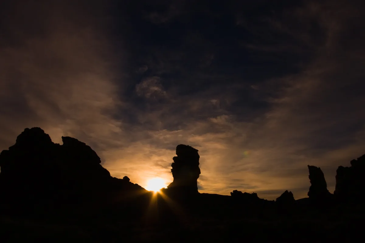 The sun setting behind dark volcanic rock formations in Teide National Park, under a streaked, darkening sky