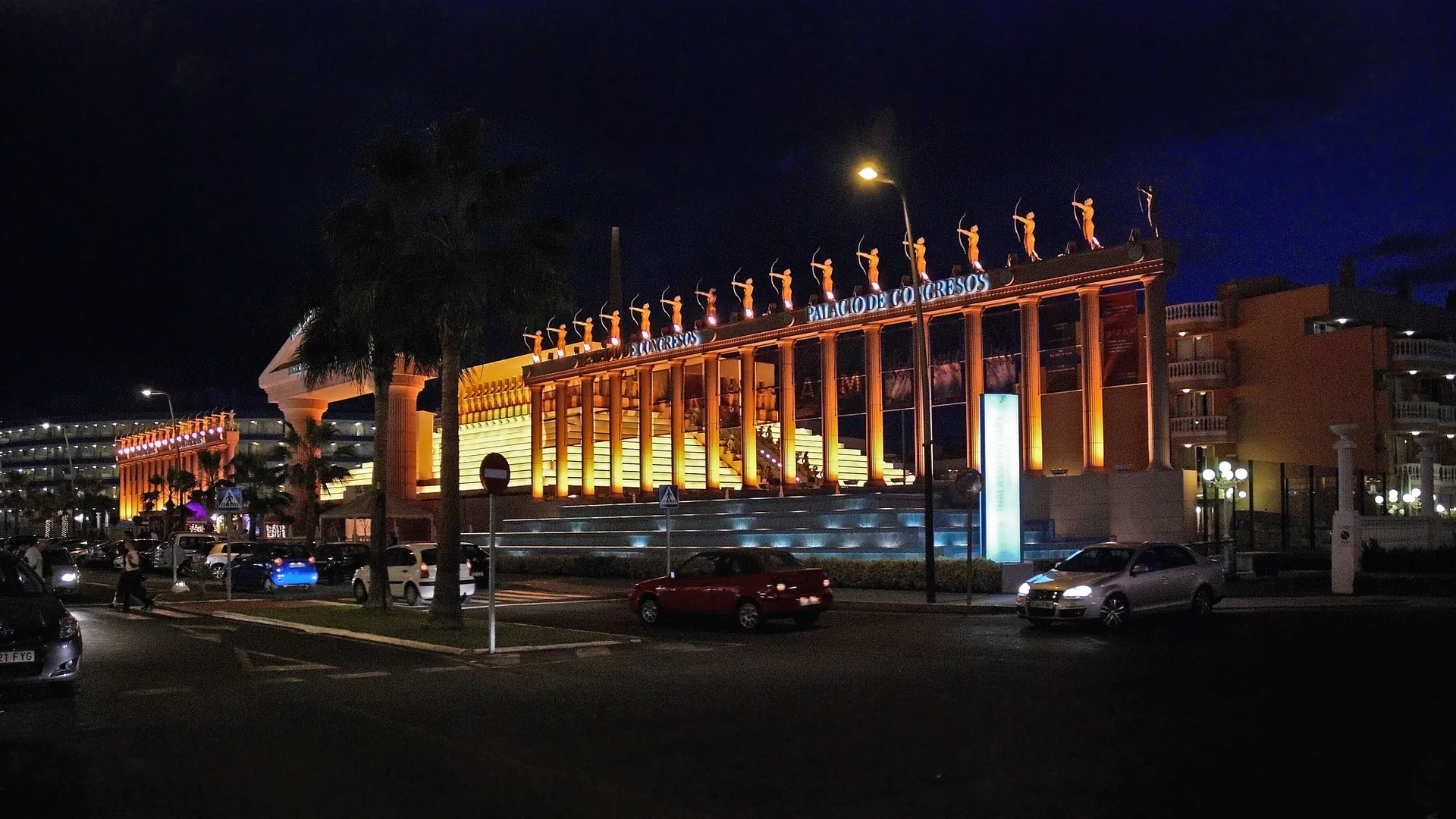 The lit colonnade of the Palacio de Congresos in Playa de las Américas at night, with golden columns, statues of archers along the roof and a deep blue sky above