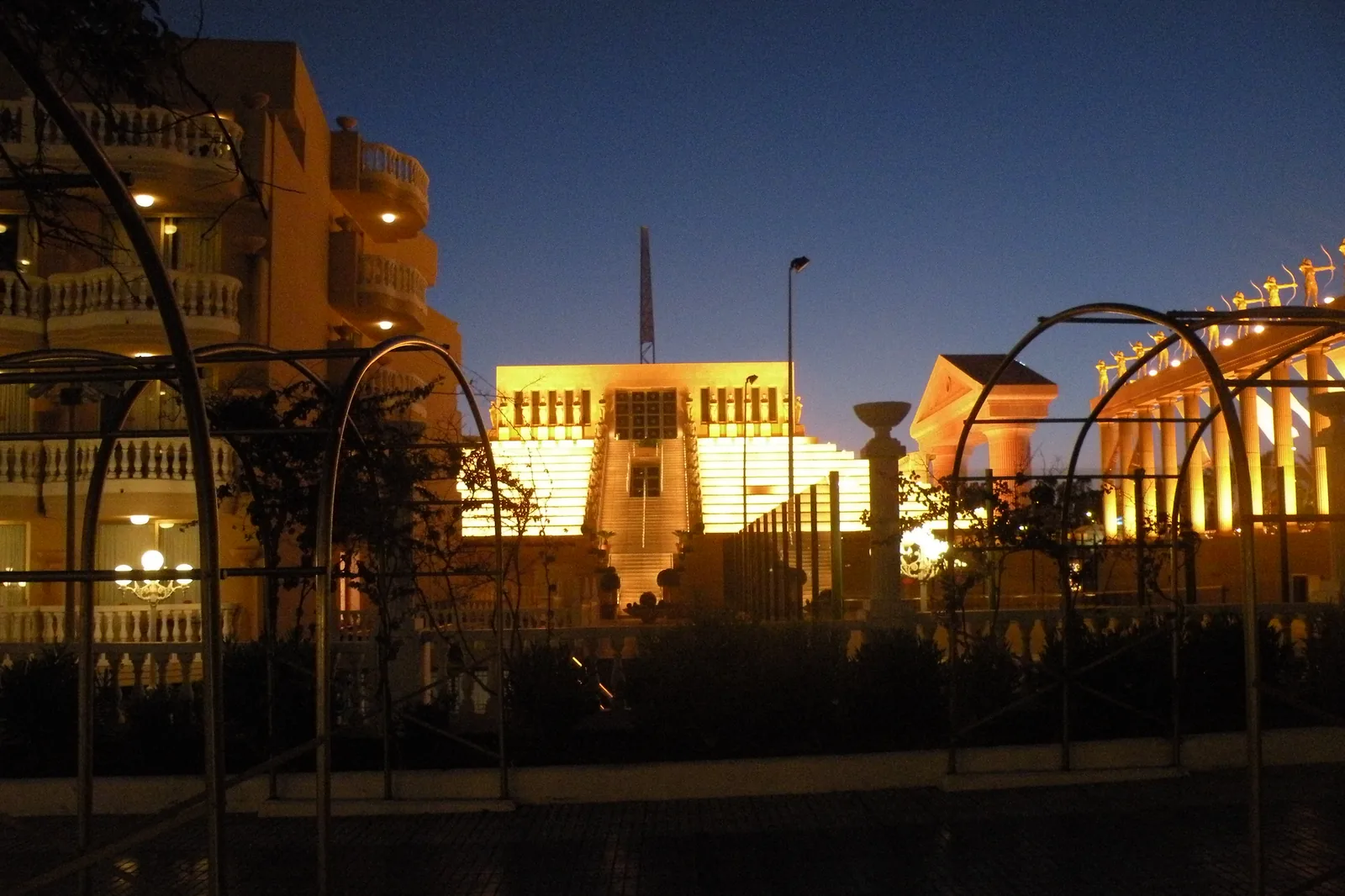 A stepped building lit gold at night beside the Palacio de Congresos colonnade, with balconies of a hotel on the left and a dark sky above